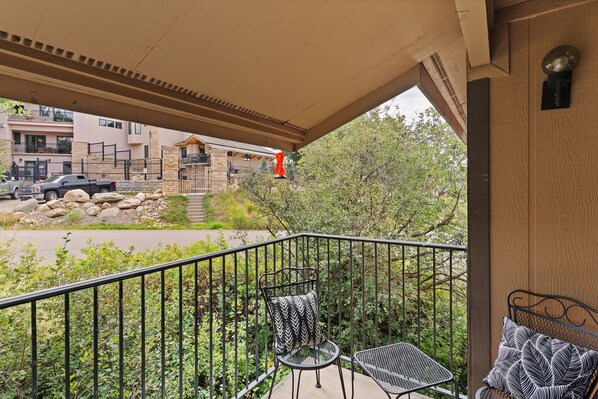 A balcony with a metal railing, a chair, and a table. There is a view of a building and a street in the background.