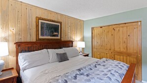 Un dormitorio con cabecera de madera, cama con ropa de cama blanca, almohada gris y un cuadro enmarcado en la pared.