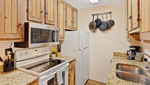 Una cocina con armarios de madera, microondas, estufa, refrigerador y una encimera de granito, además de ollas colgantes.
