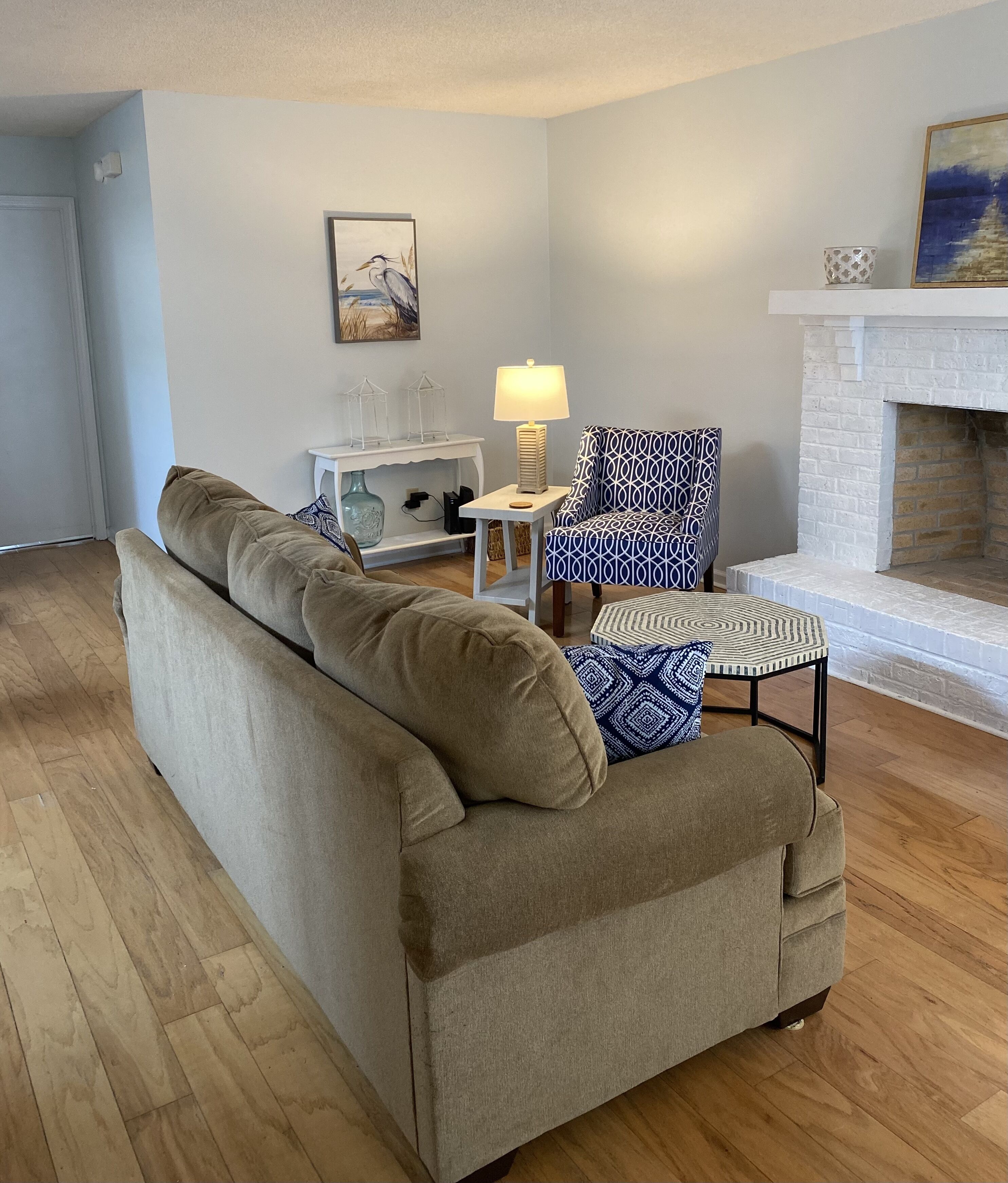 A living room with a beige sofa, a patterned armchair, a small side table with a lamp, a white fireplace, and a painting on the wall.