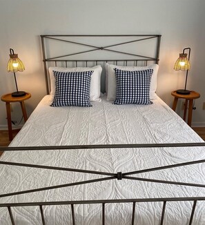 Una cama con estructura metálica, dos almohadas a cuadros y dos lámparas de noche sobre mesitas de madera.