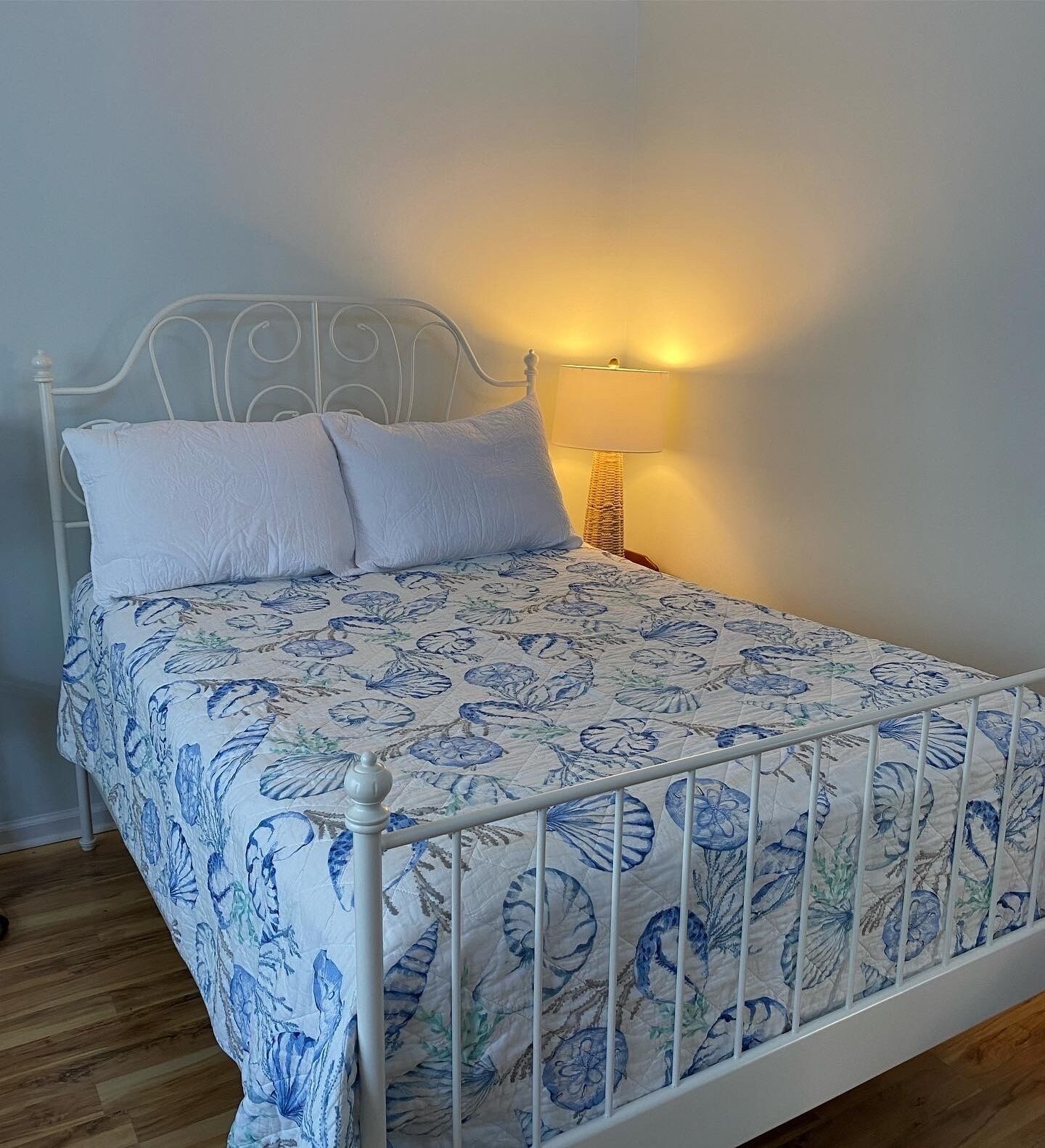 Una cama individual con colcha estampada con motivos florales, cabecera de metal blanco y lámpara de noche.