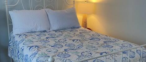 Una cama individual con colcha estampada con motivos florales, cabecera de metal blanco y lámpara de noche.