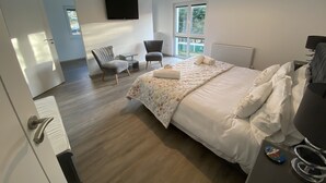 A modern hotel room with a large bed, a flat-screen TV mounted on the wall, a seating area with two chairs, and a window providing a view of the outdoors. - Sleeps8- Ecofriendly- Kingbeds- Familyfriendly (Wadebridge)