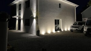 A modern two-story building with a flat roof, illuminated exterior lighting, and a driveway with parked vehicles. - Sleeps8- Ecofriendly- Kingbeds- Familyfriendly (Wadebridge)