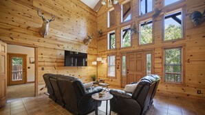 A log cabin interior with a fireplace, leather sofas, a TV, and deer antlers mounted on the wall. - PRIVATE Peaceful Cabin on 1.5 Acres with Hot Tub, Deck & HUGE Outdoor Fireplace! (Broken Bow)