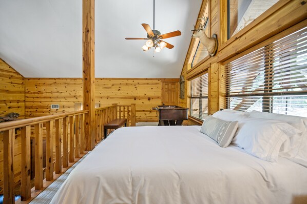 A bedroom with a wooden ceiling fan, a bed with white linens, a wooden bench, and a window with blinds.