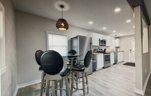 Cozinha moderna com eletrodomésticos de aço inoxidável, área de jantar com cadeiras pretas e luminária pendente.