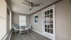 A room with a ceiling fan, a window with blinds, a white wicker chair, a small table with a basket, and a sliding glass door leading to a closet.