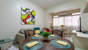 A dining area with a round wooden table set for four, surrounded by wicker chairs. A colorful abstract painting is on the wall, and there is a window with sheer curtains in the background. - 2 Minutes From Copacabana Beach A303 (Rio de Janeiro)
