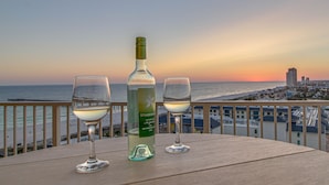 Una botella de Starborough Sauvignon Blanc y dos copas de vino en un balcón con vista a una playa y un paisaje urbano al atardecer.