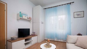 Ett vardagsrum med en tv på ett träställ, en vit soffa, ett litet runt bord med en dekorativ prydnad och ett fönster med tunna, genomskinliga gardiner.