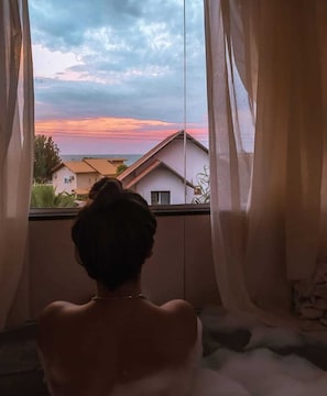En person sidder ved et vindue med lette gardiner og ser ud over et boligområde med huse i solnedgang.