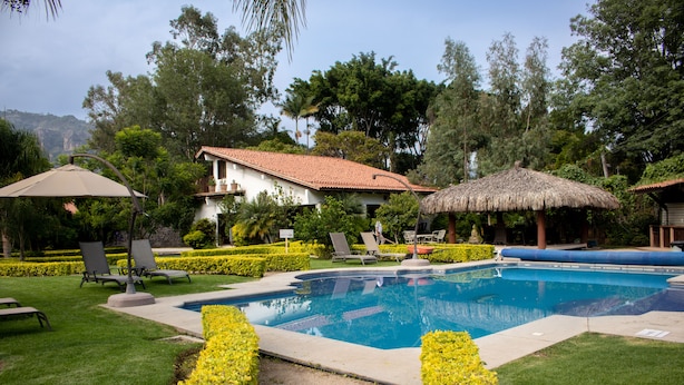 A swimming pool surrounded by neatly trimmed hedges, lounge chairs, and a thatched-roof structure. - Hotel Posada La Presa (Tepoztlán)