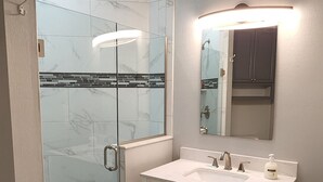 Un baño con ducha de vidrio, tocador blanco con espejo y salpicadero de azulejos.