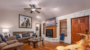 A cozy living room with a fireplace, stone walls, and a ceiling fan. - Lakefront! Hot Tub-Fire Pit, game room, paddle boards & canoe! (Kimberling City)