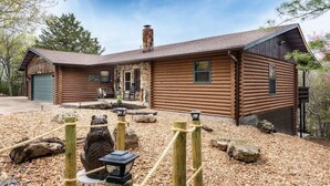 A log cabin with a stone chimney, a covered porch, and a fenced outdoor area with a bear statue. - Lakefront! Hot Tub-Fire Pit, game room, paddle boards & canoe! (Kimberling City)