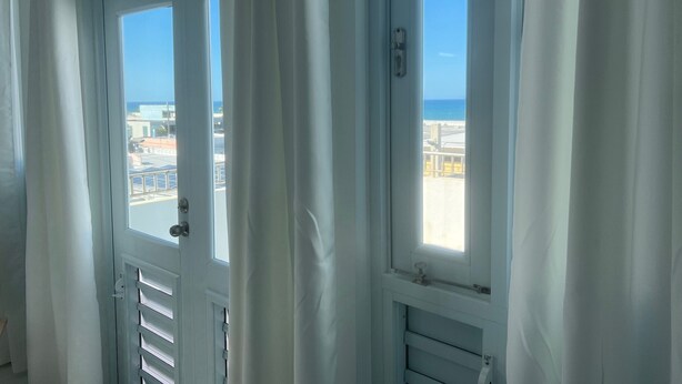 A room with a view of the beach and buildings through a window with white curtains. - Arecibo Hotel 4301 (Arecibo)