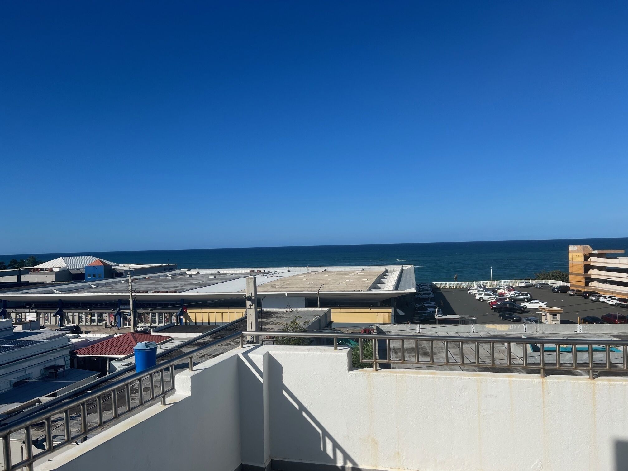 A rooftop view with a clear blue sky, a parking area, and a distant view of the ocean.