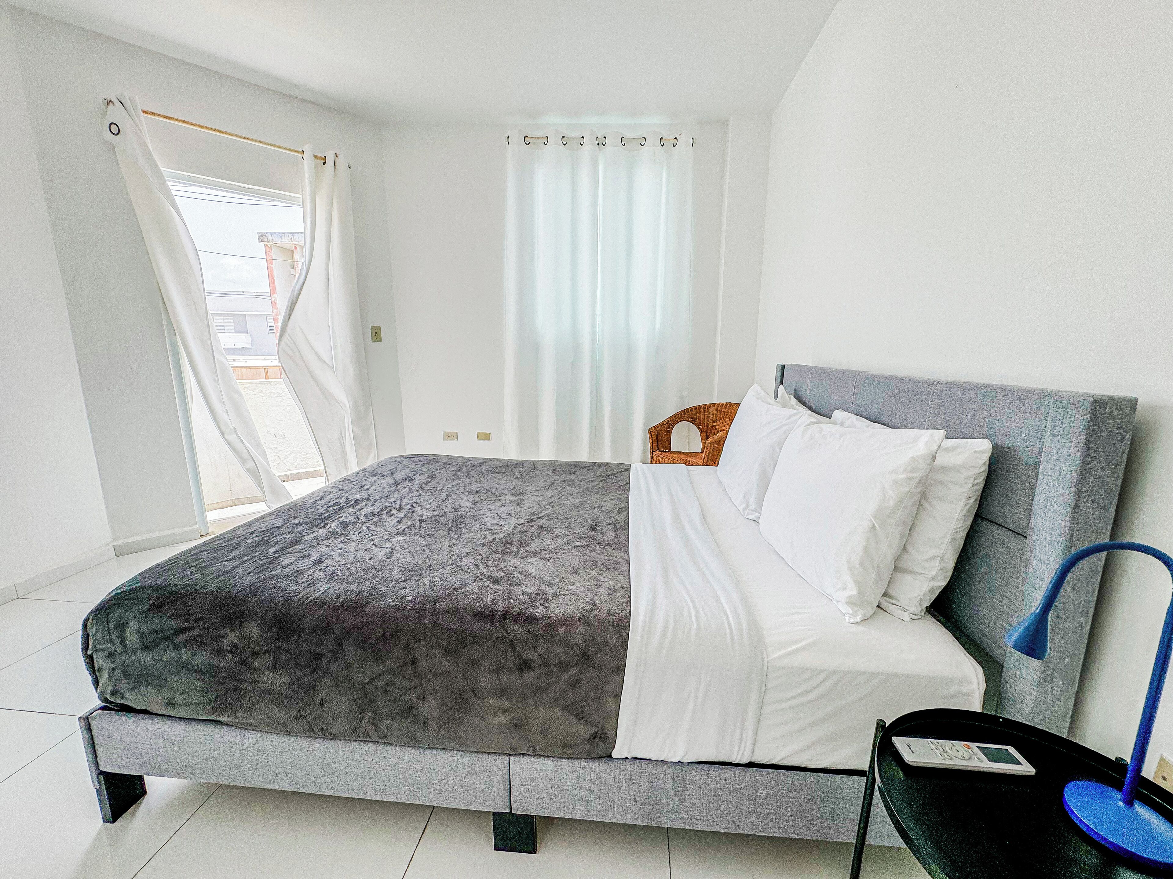 A neatly made bed with white linens, a grey headboard, and a bedside table with a lamp.