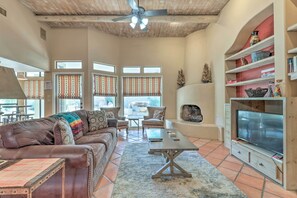 A spacious living room with a brown leather sofa, a coffee table, and a TV on a wooden stand. There are windows with curtains, a ceiling fan, and a bookshelf with decorative items. - 6 Mi to Dtwn Mesa! Pueblo Retreat w/ Patio (Mesa)