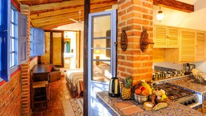 A cozy room with a wooden ceiling, a brick wall, a bed, a table with a kettle, and a kitchen area with a stove and various food items. - Casa Aurora Garden Retreat (Ajijic)