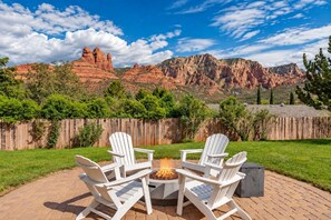 A patio with Adirondack chairs and a fire pit, surrounded by a wooden fence and a view of red rock formations. - Southwest uptown retreat w/Panoramic Views/ Hottub (Sedona)