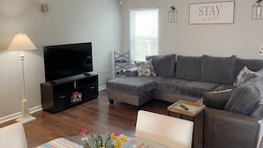 A modern living room with a sectional sofa, a flat-screen TV, a dining table with a floral centerpiece, and a wall with a 'STAY while you wait' sign.