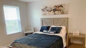 A bedroom with a bed, a chair, a nightstand, a window, and a painting on the wall.
