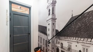 A hallway with a door, a trash bin, and a mural of a building with a clock tower. - Coimbra City Charm (Coimbra)