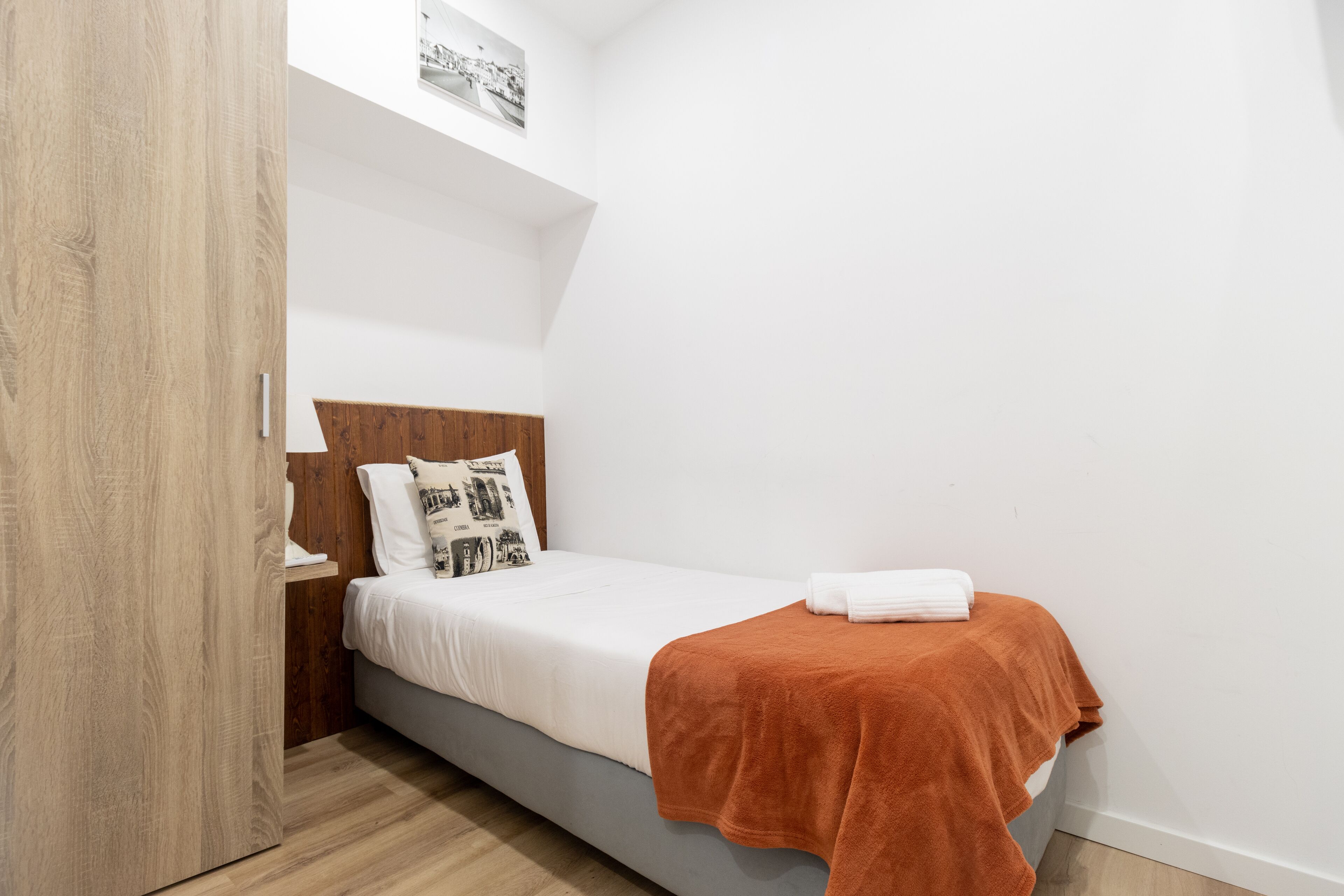 Un dormitorio pequeño y ordenado con una cama individual, un armario de madera y una pared blanca con un cuadro.