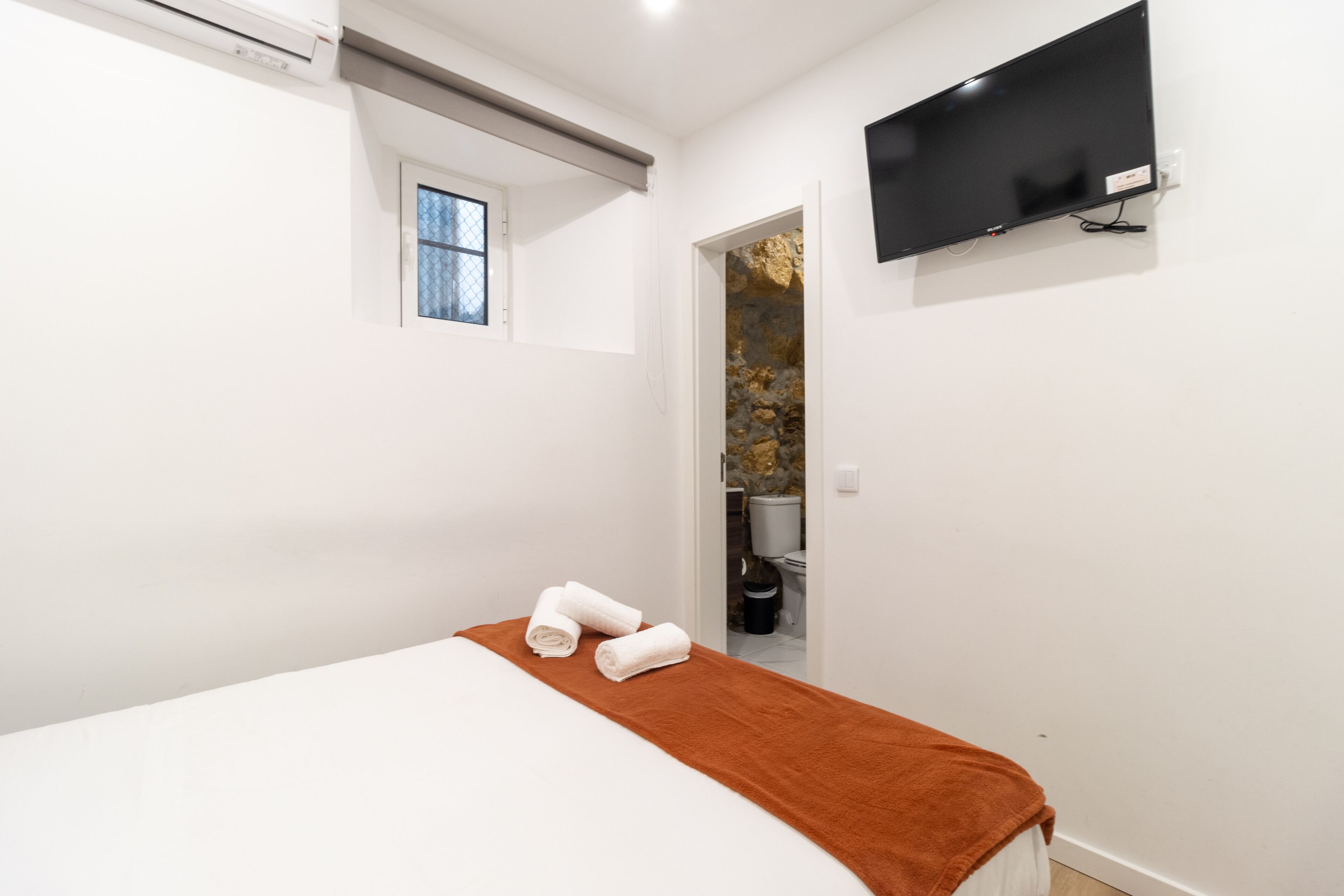 Un dormitorio con una cama, un televisor montado en la pared, una ventana y una puerta que da a un baño.