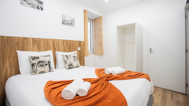 A hotel room with a wooden headboard, two white beds with matching pillows, an orange throw blanket, and a wooden door. - Coimbra City Charm (Coimbra)