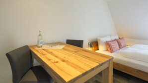 A room with a wooden table, a chair, a bed with pillows, and a bedside table with a lamp.