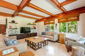Un salon spacieux avec un plafond haut, des poutres en bois, un téléviseur, un canapé, une table basse et un fauteuil en osier.