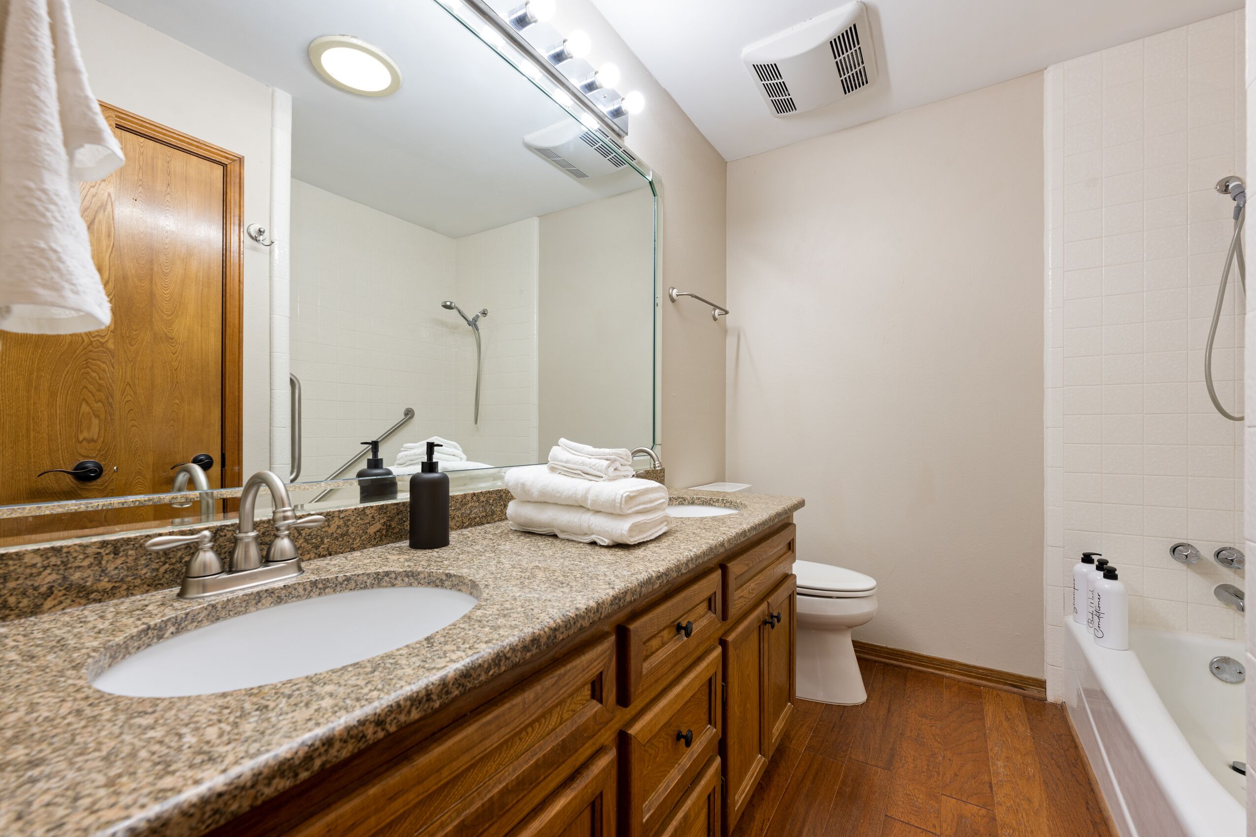 Une salle de bain avec un lavabo sur un plan de travail en granit, un meuble-lavabo en bois, un miroir, des toilettes et une baignoire.