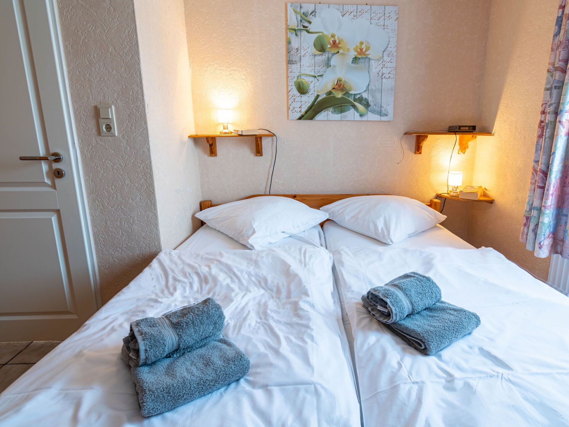 Ein ordentlich bezogenes Bett mit weißen Leinen und zwei gefalteten Handtüchern, zwei an der Wand befestigten Leselampen und einem floralen Kunstwerk darüber.