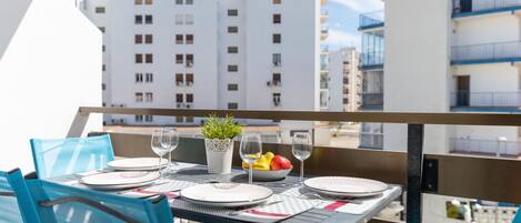 A balcony with a table set for two, featuring a potted plant, fruit bowl, and wine glasses.