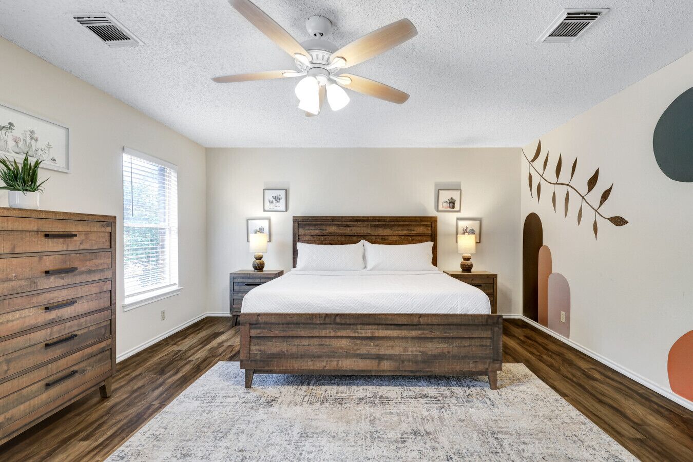 A bedroom with a wooden bed, a ceiling fan, a dresser, and a wall with abstract art.