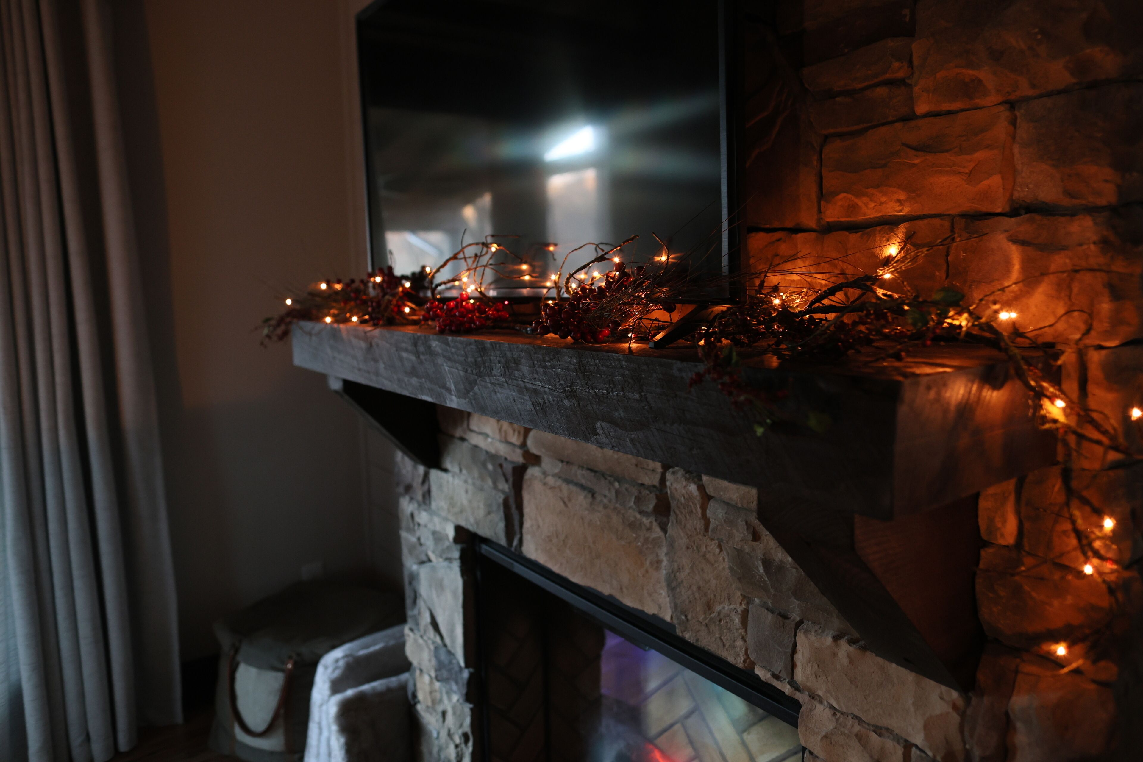 A stone fireplace with a flat-screen TV mounted above it, decorated with string lights and red berries.