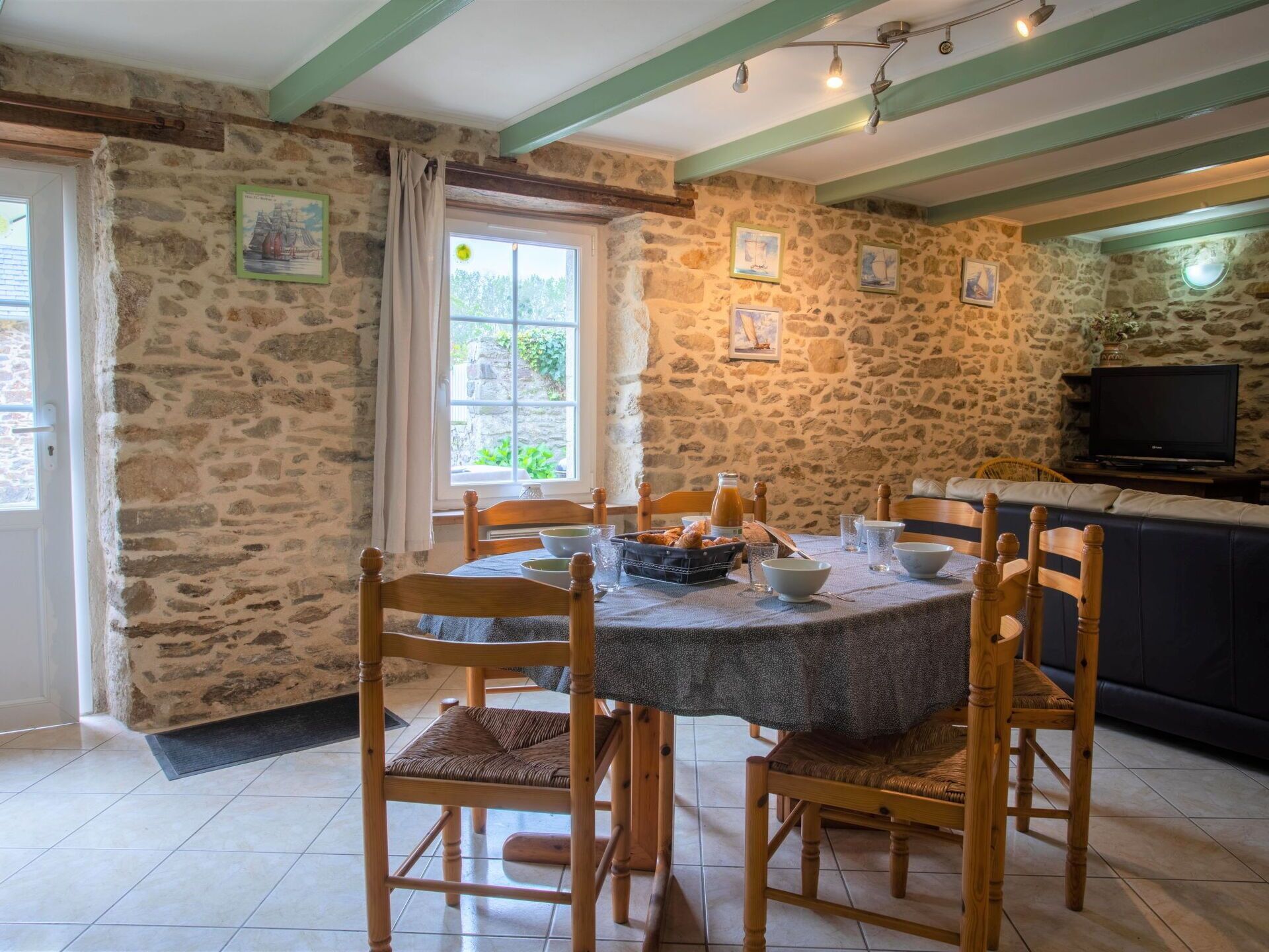 Un coin repas aux murs de pierre, avec une table ronde, des chaises en bois et un téléviseur en arrière-plan.