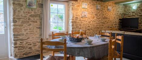 Un coin repas aux murs de pierre, avec une table ronde, des chaises en bois et un téléviseur en arrière-plan.
