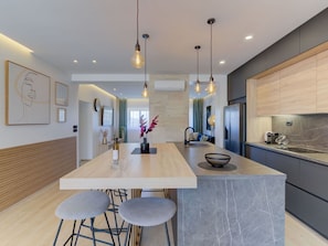 A modern kitchen with a central island, bar stools, pendant lighting, and a view into the living area.