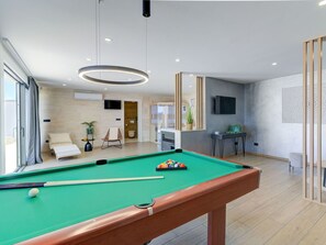 A modern indoor pool area with a pool table, a lounge chair, and a television mounted on the wall.
