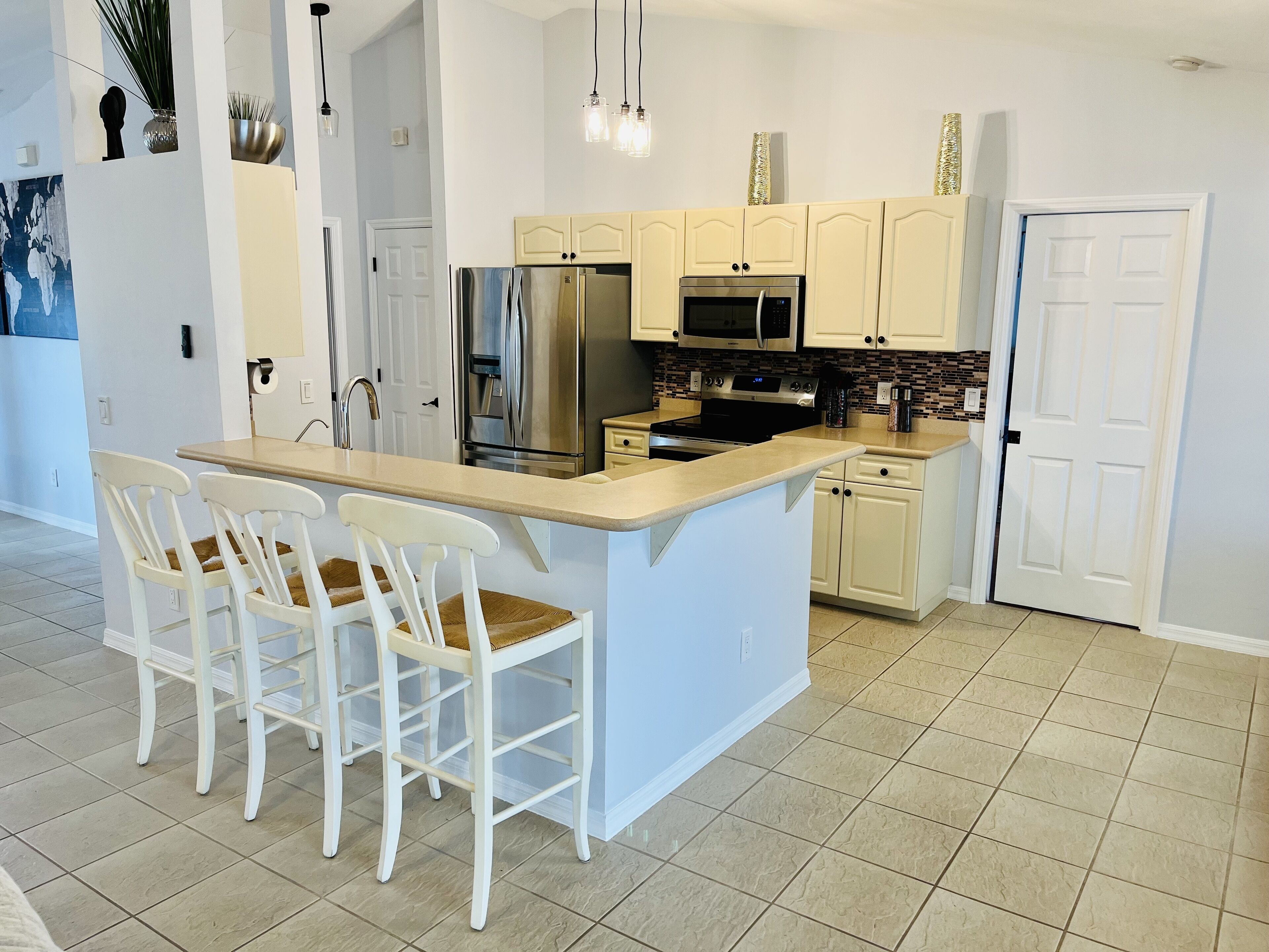 A kitchen with a central island, white bar stools, stainless steel appliances, and a tiled floor.