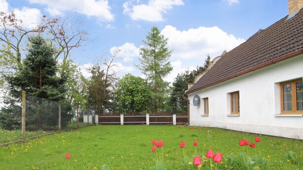 Une maison blanche avec un toit brun, entourée d’une pelouse verte parsemée de fleurs rouges et jaunes.