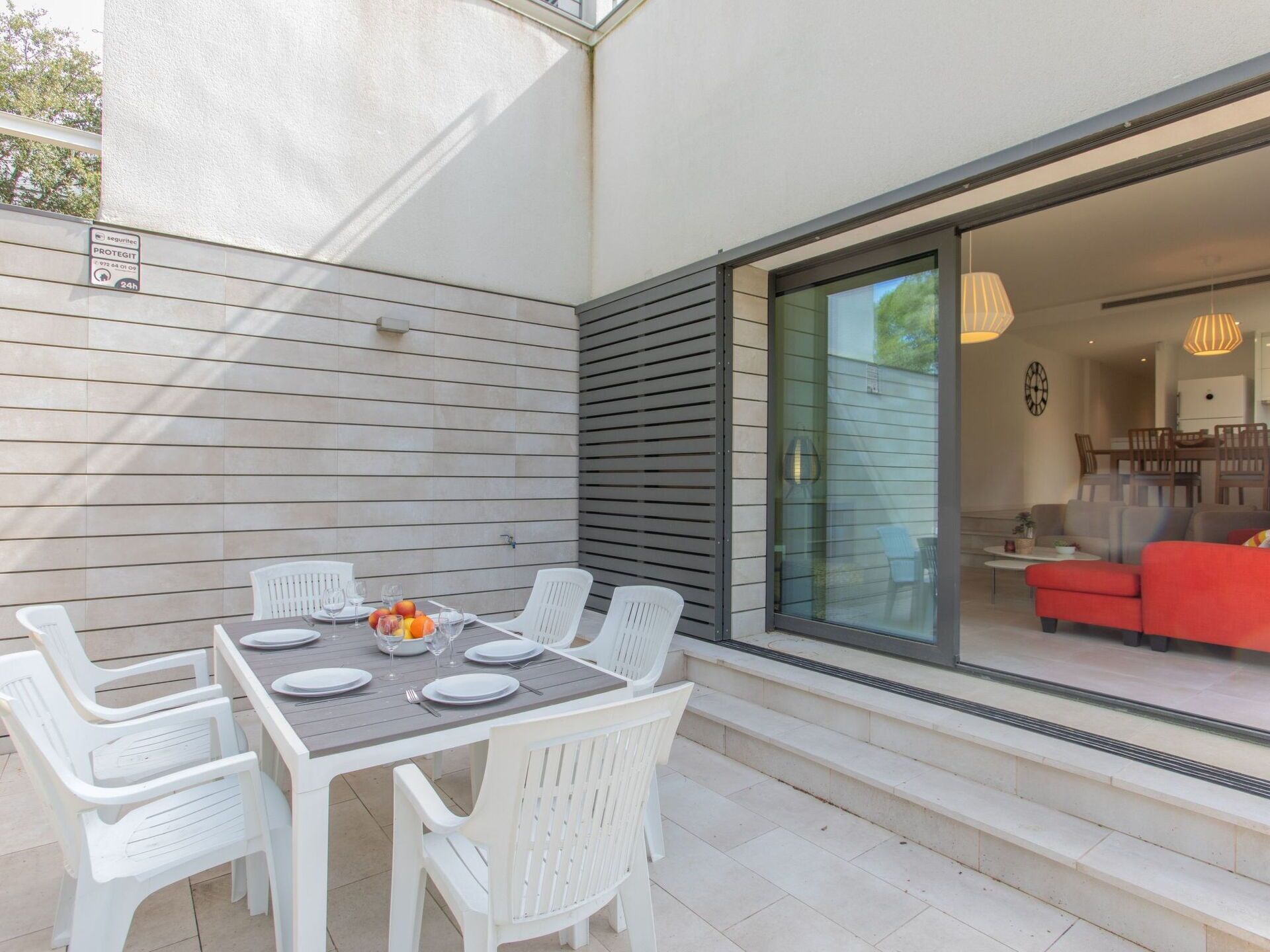 An outdoor dining area with a table set for four, surrounded by white chairs, and a glass door leading to a living room with a red sofa.