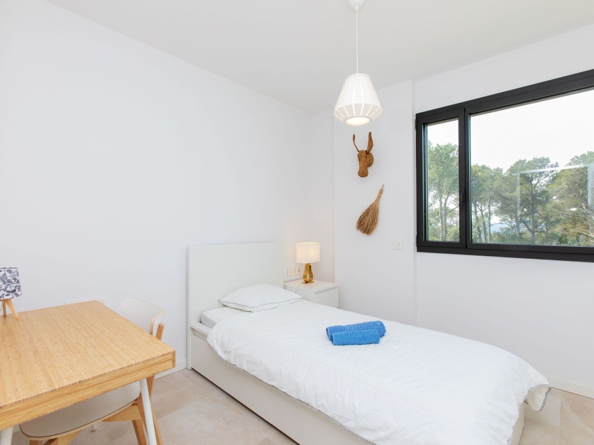 Ein schlicht eingerichtetes Schlafzimmer mit Bett, Nachttisch, Lampe und einem Fenster mit Blick auf Bäume.