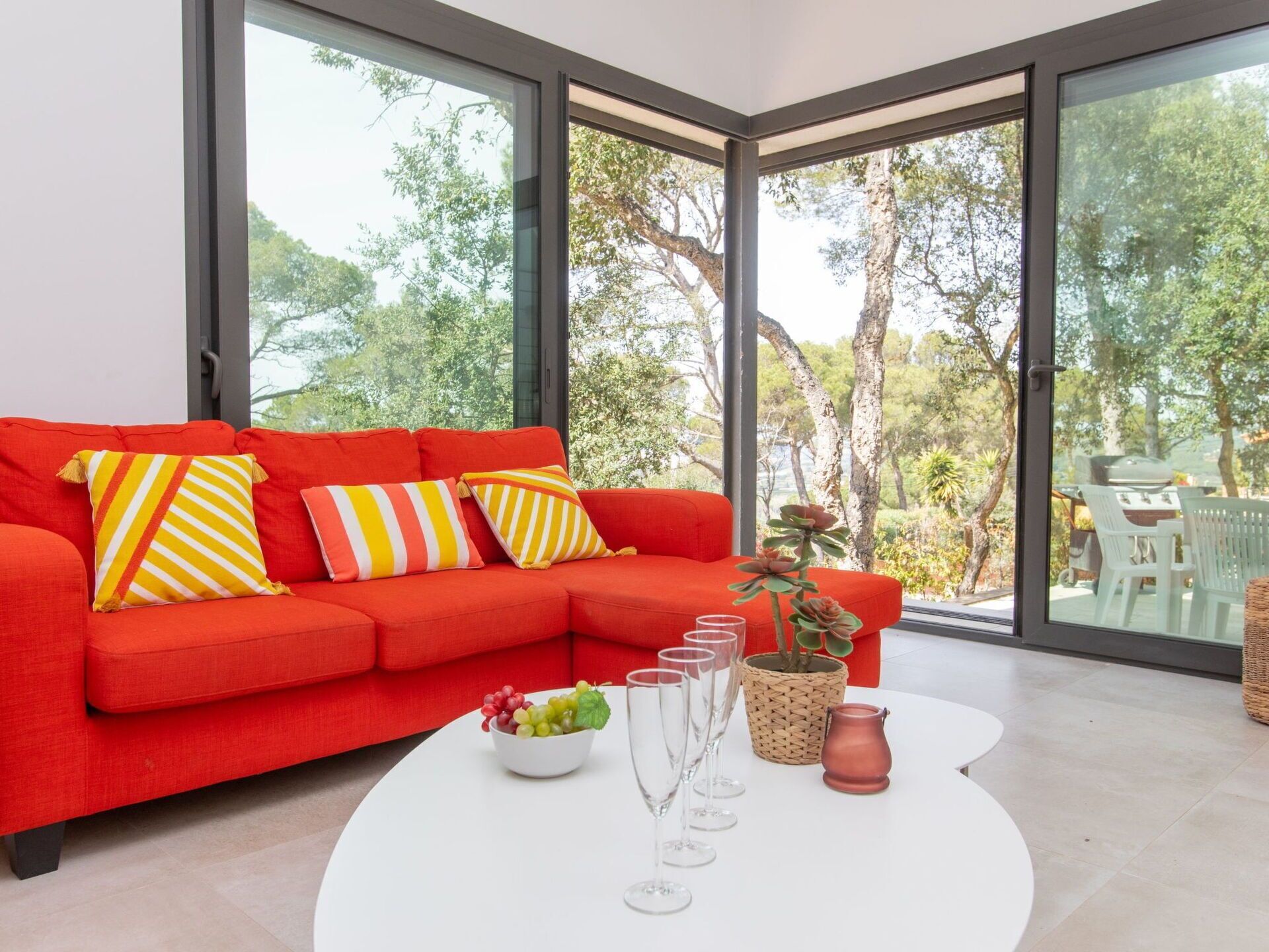 A bright red sofa with striped pillows, a white coffee table with a bowl of fruit and wine glasses, and a view of a garden through large windows.