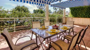 An outdoor patio with a table set for dining, surrounded by chairs, under striped awnings, with a view of greenery and buildings in the background.